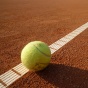 Brown ground of a tennis court with a white boundary line. A yellow tennis ball sits on the white boundary line. 