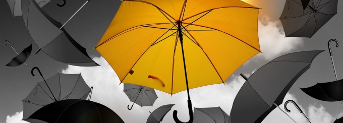 Black and white image of umbrellas floating in the sky. The center umbrella is yellow. 