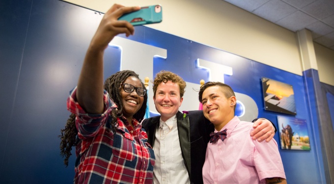 Three students pose for a selfie in front of the UB logo. 