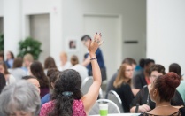 dark haired female student at table raising hand. 