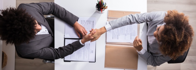 Two people sitting at a table across from each other. They are shaking hands after a job interview. 