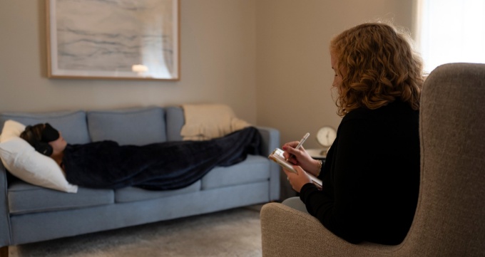 Glynn Couch seated across from a client who is lying on a sofa, wrapped in a blanket, wearing a head and eye covering, while ketamine is administered.