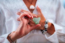 Woman wearing a white linen shirt holding one hand to her heart while her other hand holds out a crystal. 